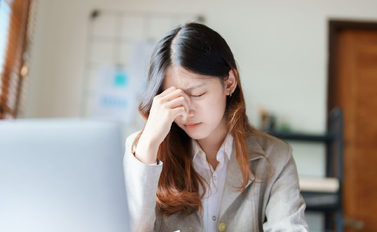 這張圖片顯示一位女性坐在桌前，手揉著鼻梁，眼睛閉著，顯示出她感覺眼有異物感的不適。這圖片適合用於討論長時間使用電腦或工作壓力引起的眼部疲勞和異物感，並提醒人們正確護理眼睛的重要性。