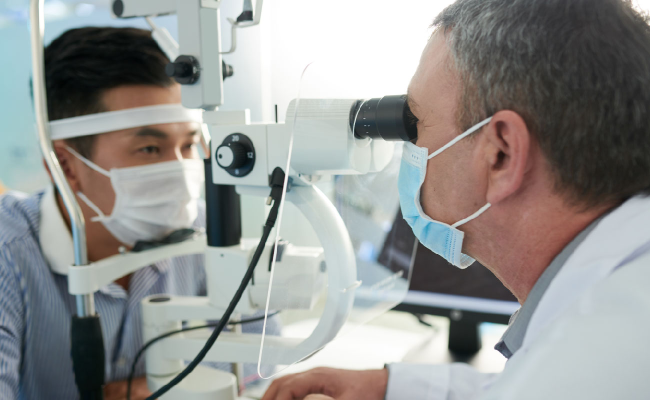 這張圖片顯示一位醫生正在使用裂隙燈顯微鏡檢查一位患者的眼睛。兩人都戴著口罩，強調了醫療環境的安全防護措施。這場景適合用於討論眼科檢查的重要性，尤其是當患者感覺眼有異物感時，專業檢查可以幫助確定問題並提供適當的治療建議。
